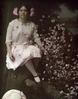 Woman in a pink dress posed in a chair in a garden scene hat off